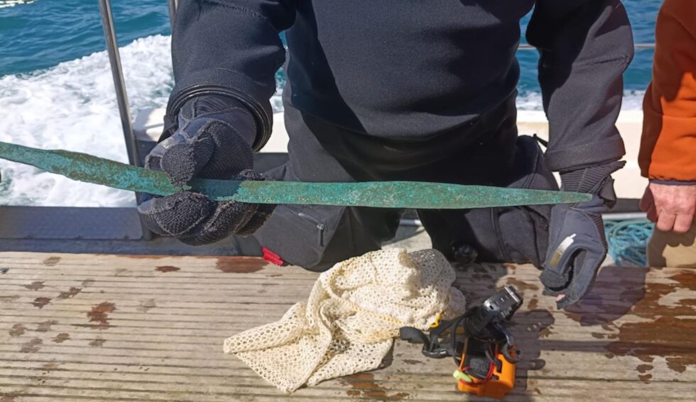 A person holding a long, rusted metal sword on a boat with water in the background.