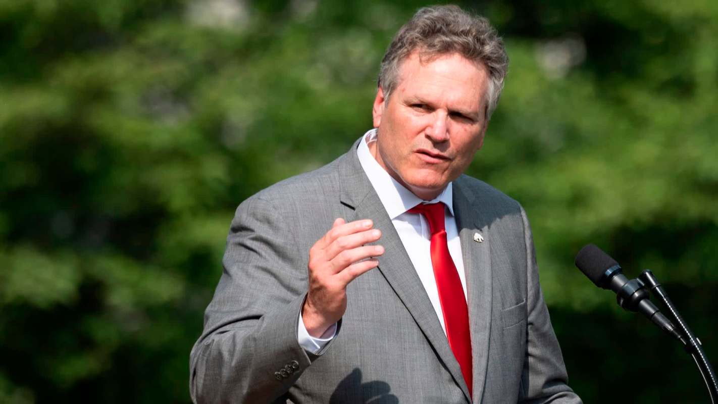 Alaska governor Mike Dunleavy in a suit speaks at a podium outdoors with greenery in the background.