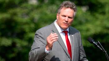 Alaska governor Mike Dunleavy in a suit speaks at a podium outdoors with greenery in the background.