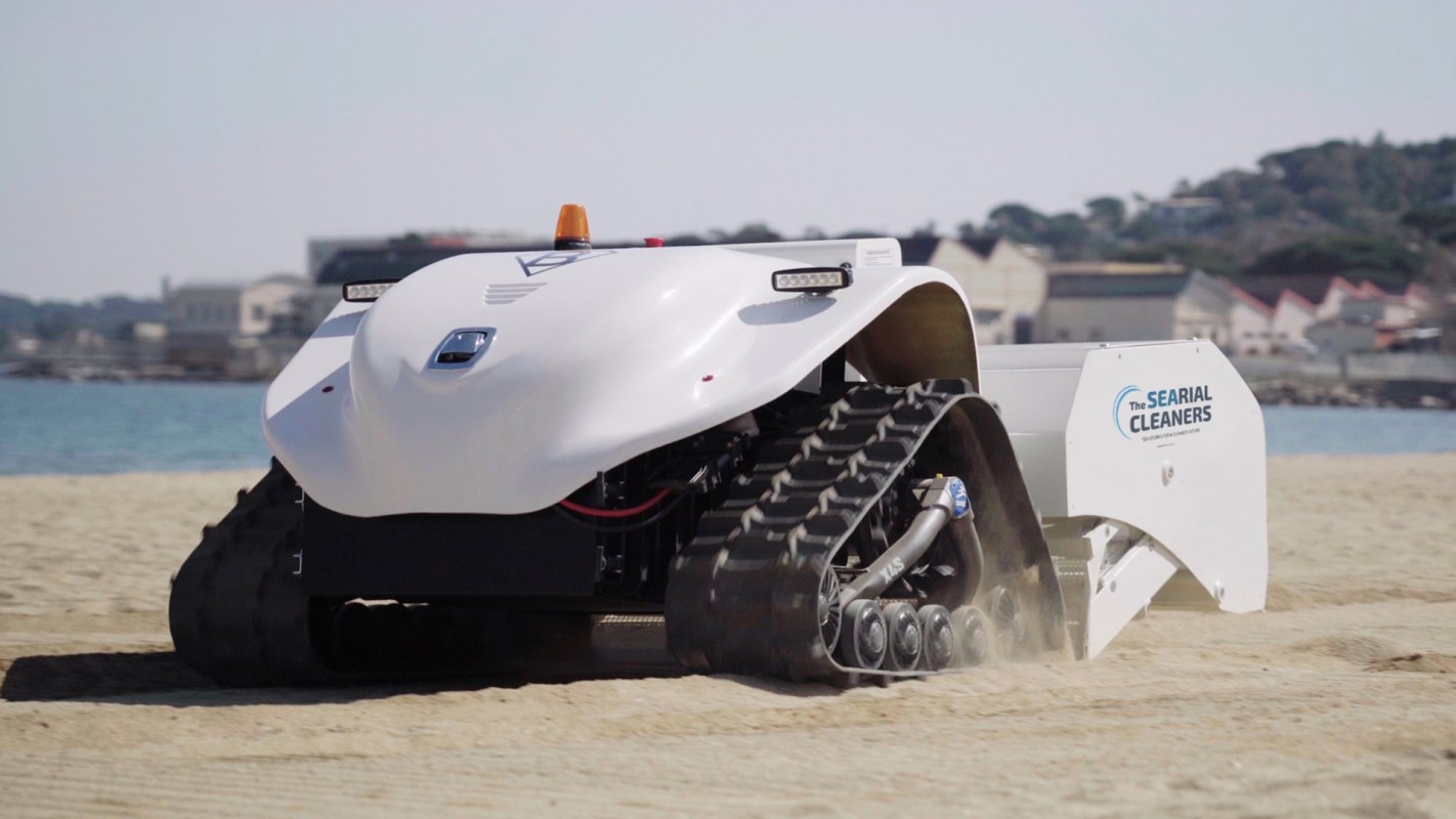 A robotic beach cleaner operates on sandy terrain near the water.