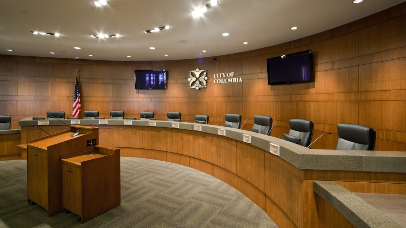 A meeting room with a curved table, several chairs, and a podium, featuring a city emblem and an American flag.