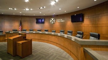 A meeting room with a curved table, several chairs, and a podium, featuring a city emblem and an American flag.