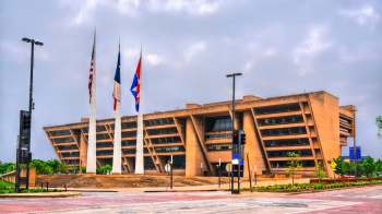 Dallas City Hall with large windows, flanked by several flags, and surrounded by landscaping and roadways.