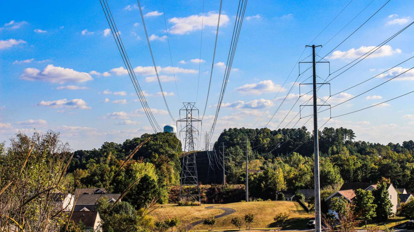 Power lines extend over a grassy area with houses and trees in the background under a blue sky with clouds.