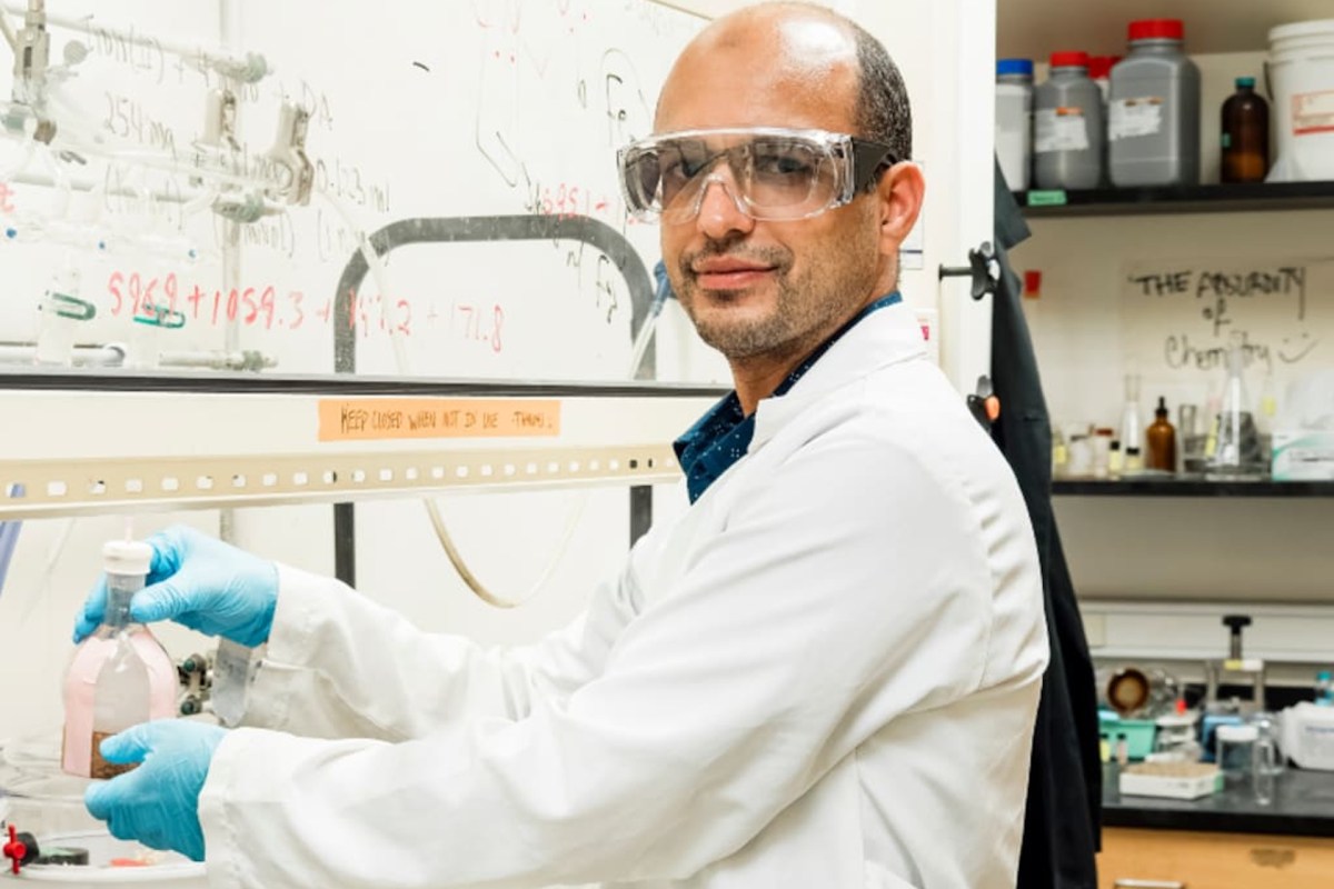A scientist in a laboratory holds a bottle while standing near a whiteboard with calculations.
