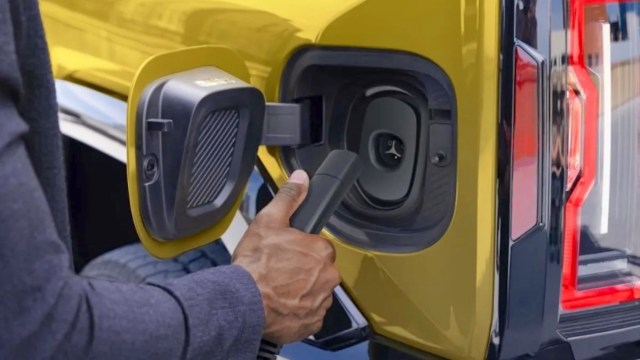 A hand holds an electric vehicle charging connector near a charging port on a yellow vehicle.