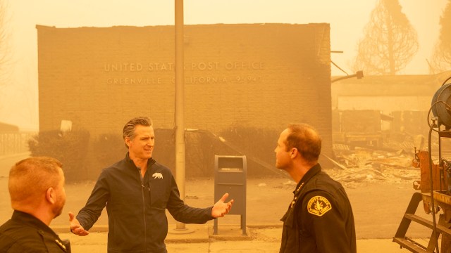 California Gov. Gavin Newsom gestures to two people near a post office in a smoky, burned area with visible debris,
