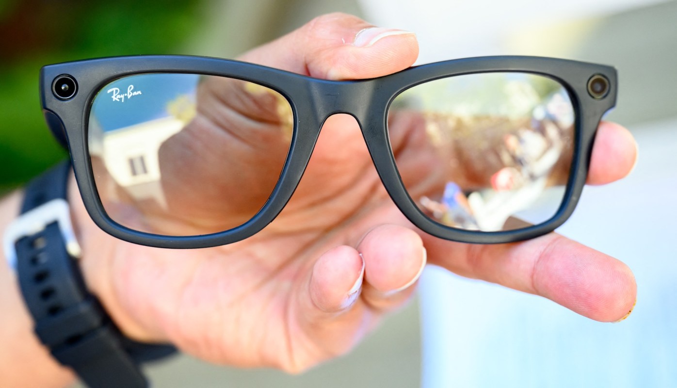 A hand holds a pair of black Meta Ray-Ban sunglasses reflecting a colorful scene.