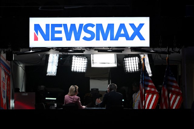 A Newsmax studio with two hosts seated at a table, illuminated by studio lights and American flags in the background.