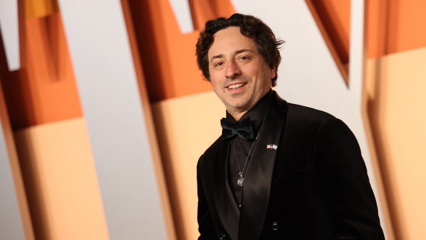 Google co-founder Sergey Brin, in a black tuxedo with a bow tie, stands against a colorful backdrop.