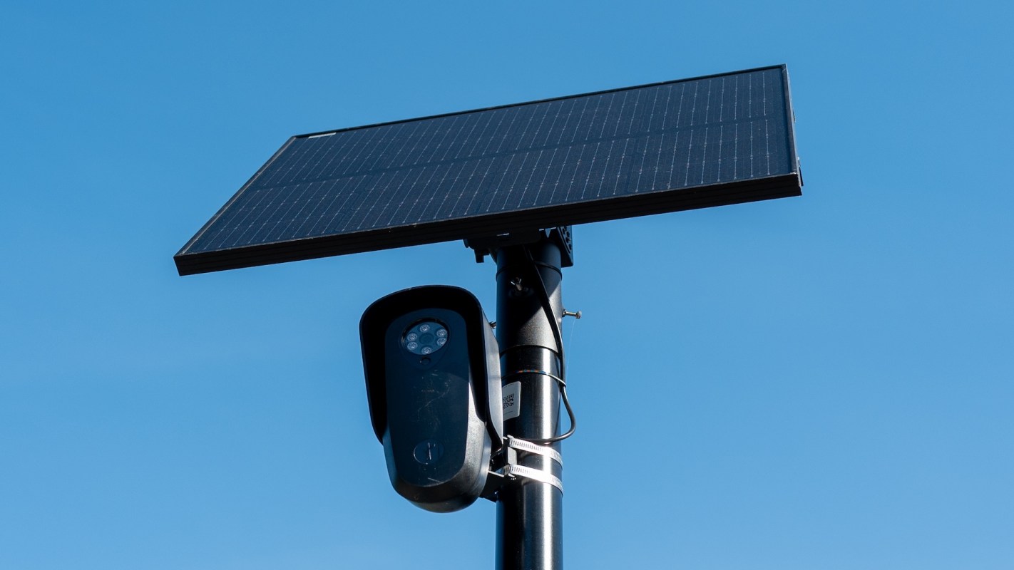 A solar panel mounted on a pole with a camera underneath against a clear blue sky.