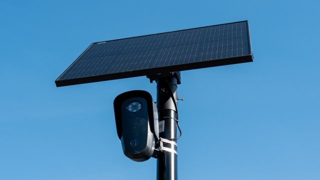 A solar panel mounted on a pole with a camera underneath against a clear blue sky.