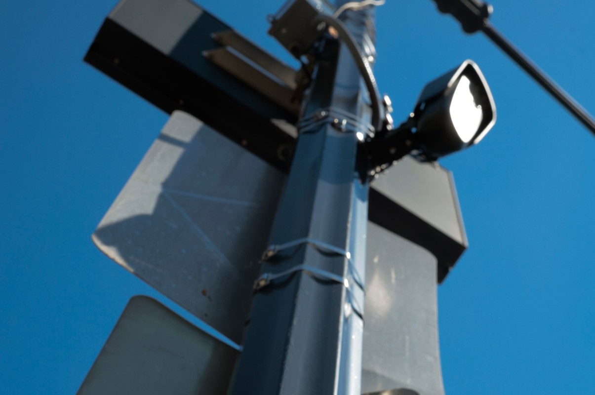 A Flock camera and a traffic signal mounted on a pole with a spotlight and blue sky in the background.