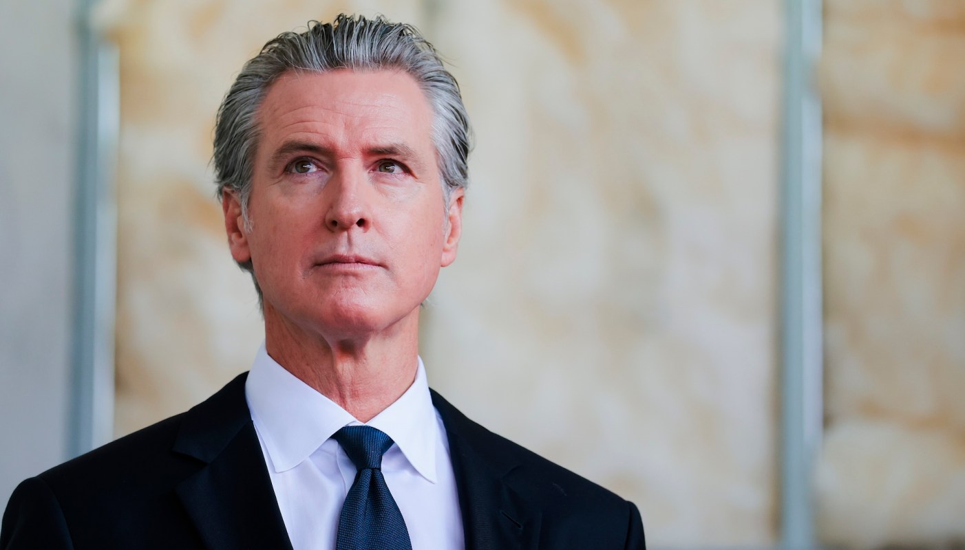 Gov. Gavin Newsom, wearing a suit, stands in an indoor setting with a neutral background.