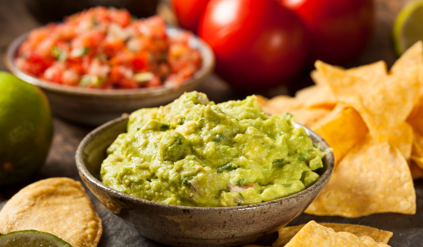 A bowl of guacamole served with tortilla chips, lime, and a bowl of salsa in the background.