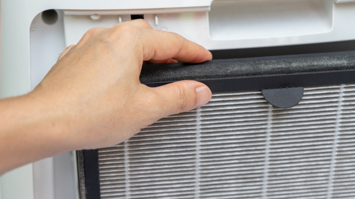 A hand holds a filter near an air conditioning unit.