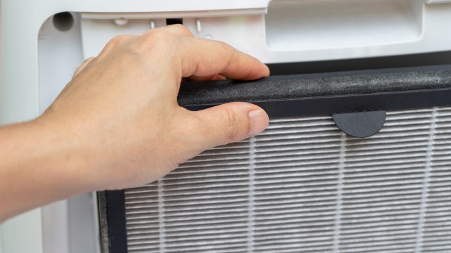 A hand holds a filter near an air conditioning unit.