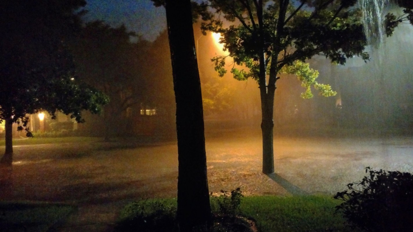 A heavily rain-soaked street illuminated by nearby lights and surrounded by trees.