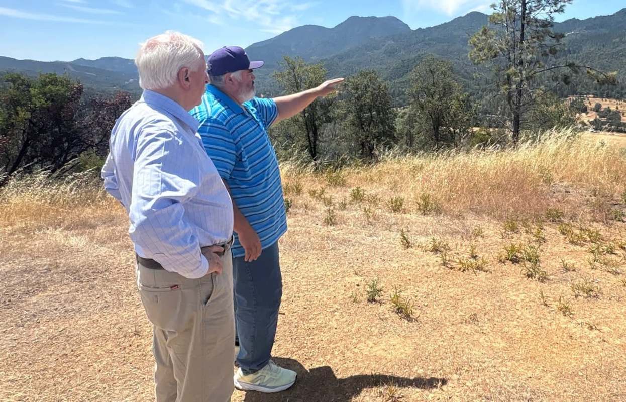 Two men stand outdoors, one pointing toward a mountainous landscape in the background.