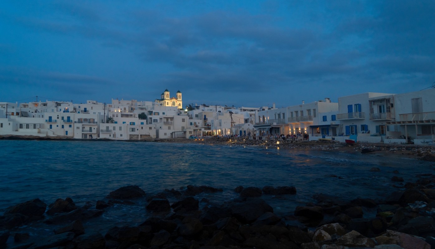 A coastal scene featuring white buildings along the shore under a twilight sky, with a church in the background.