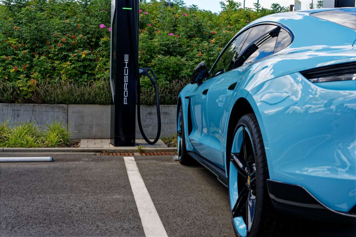 A light blue Porsche electric vehicle charging at a station surrounded by green foliage.