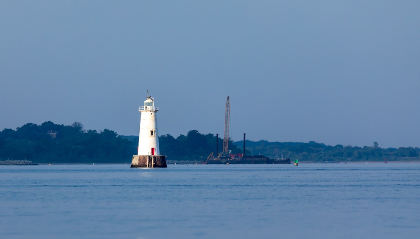 A lighthouse stands in the water with a construction barge in the background.
