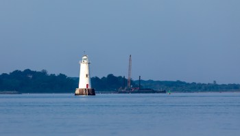 A lighthouse stands in the water with a construction barge in the background.