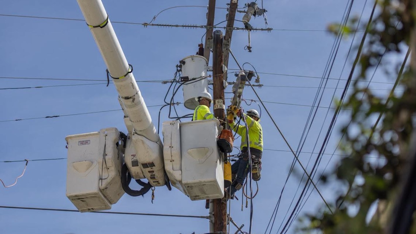 Two workers in safety gear are performing maintenance on a utility pole with power lines.