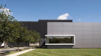 A modern building with a glass section surrounded by trees and grass under a clear blue sky.