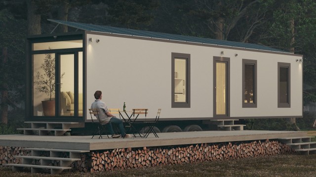 A tiny house on a wooden deck with stacked firewood, featuring large windows and solar panels, and a person sitting at a table.