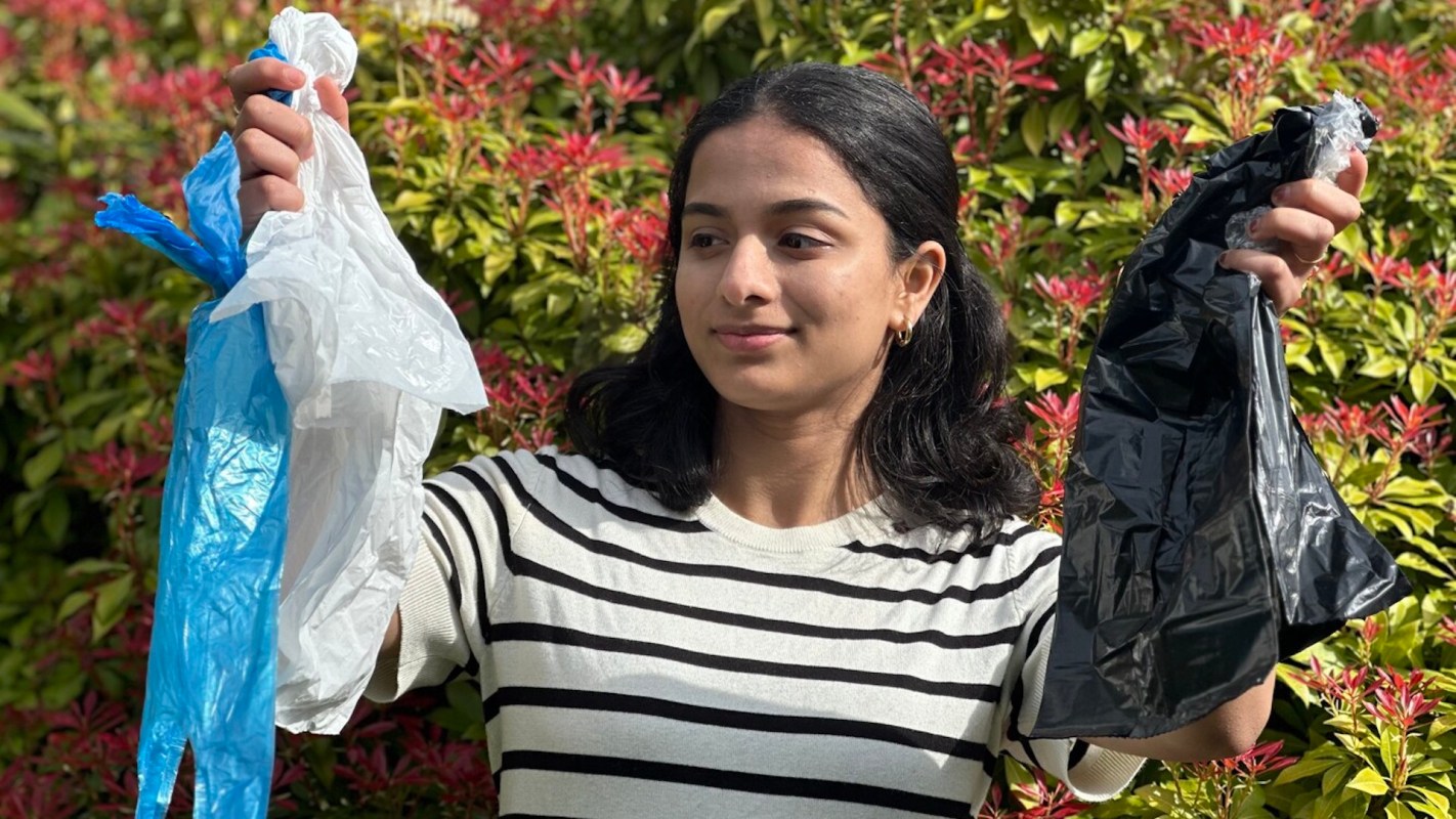 A person holds a blue plastic bag in one hand and a black plastic bag in the other, with greenery in the background.
