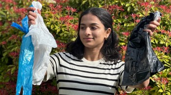 A person holds a blue plastic bag in one hand and a black plastic bag in the other, with greenery in the background.
