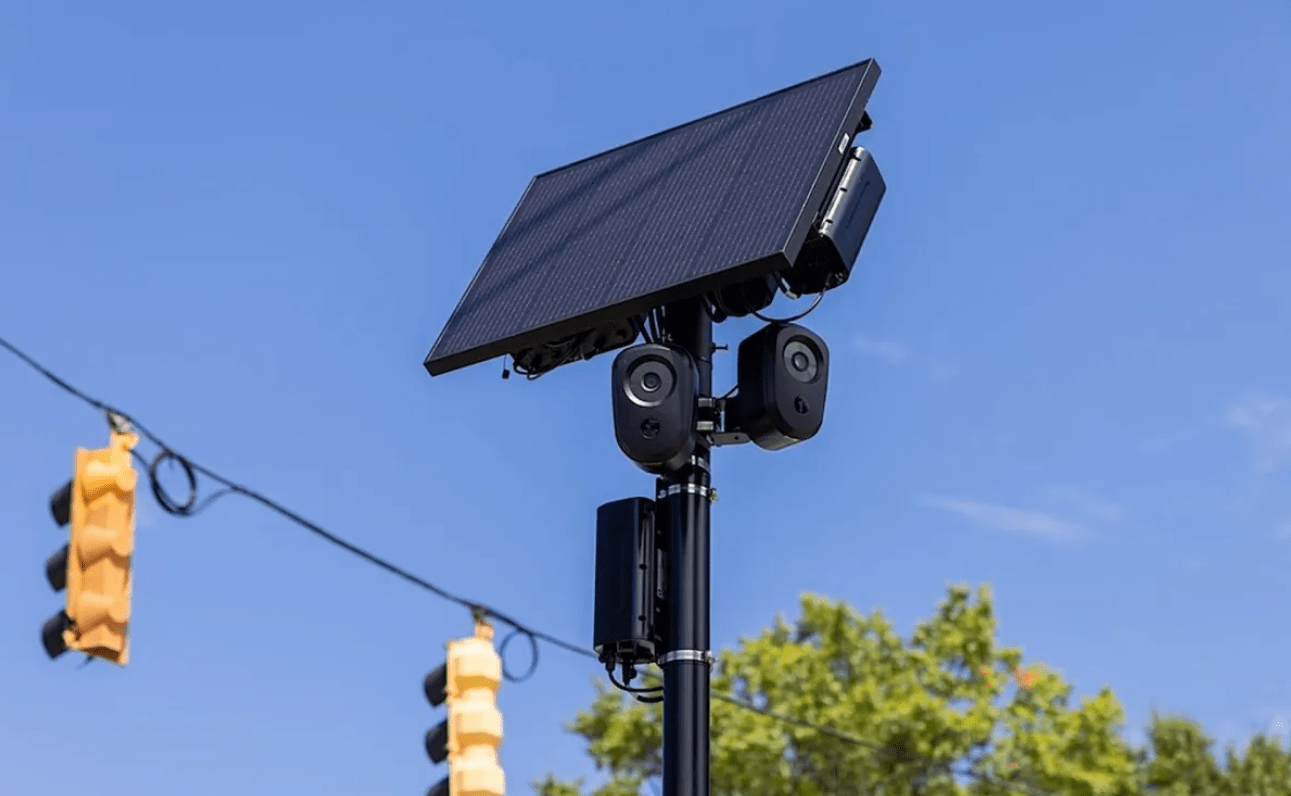 A solar panel mounted on a pole with cameras, near traffic lights and trees against a blue sky.