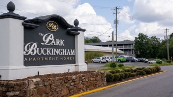 A sign for The Park Buckingham Apartment Homes with parked cars and building in the background.