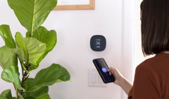 A person adjusts a smart thermostat while holding a smartphone, with a potted plant nearby.