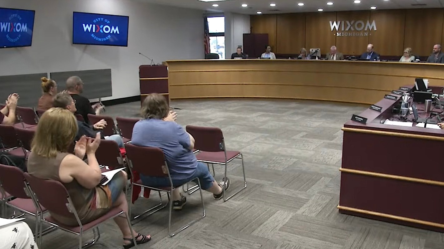 A city council meeting taking place in Wixom, Michigan, with attendees sitting in chairs and speakers at a podium.