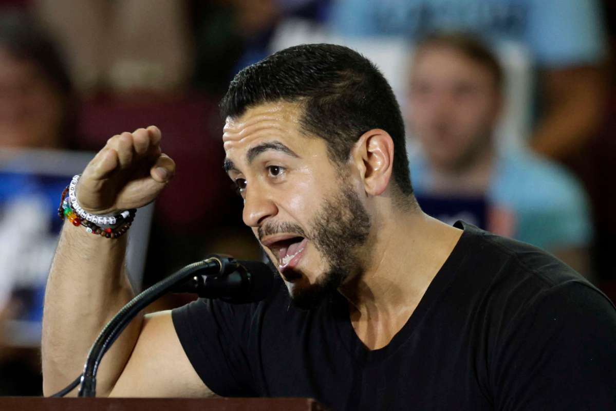 A man speaks passionately at a microphone during an event, with a blurred audience in the background.