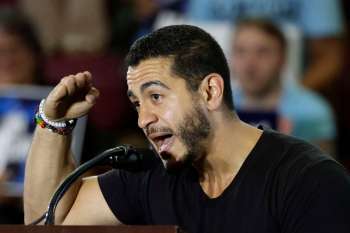 A man speaks passionately at a microphone during an event, with a blurred audience in the background.