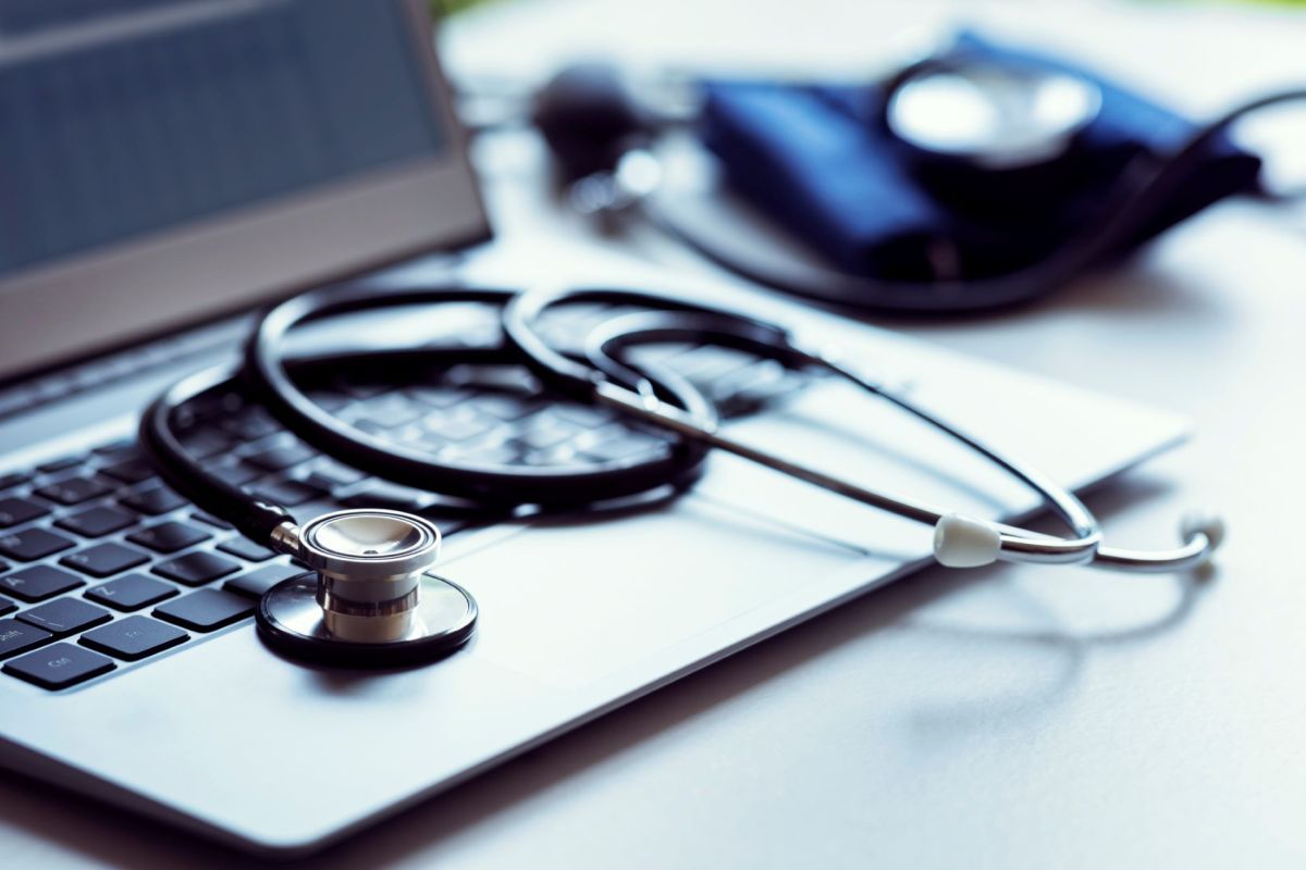 A stethoscope rests on a laptop keyboard with a blood pressure cuff in the background.