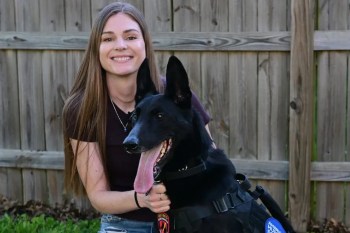 A woman poses with a black dog in a yard by a wooden fence.