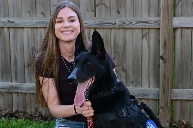 A woman poses with a black dog in a yard by a wooden fence.