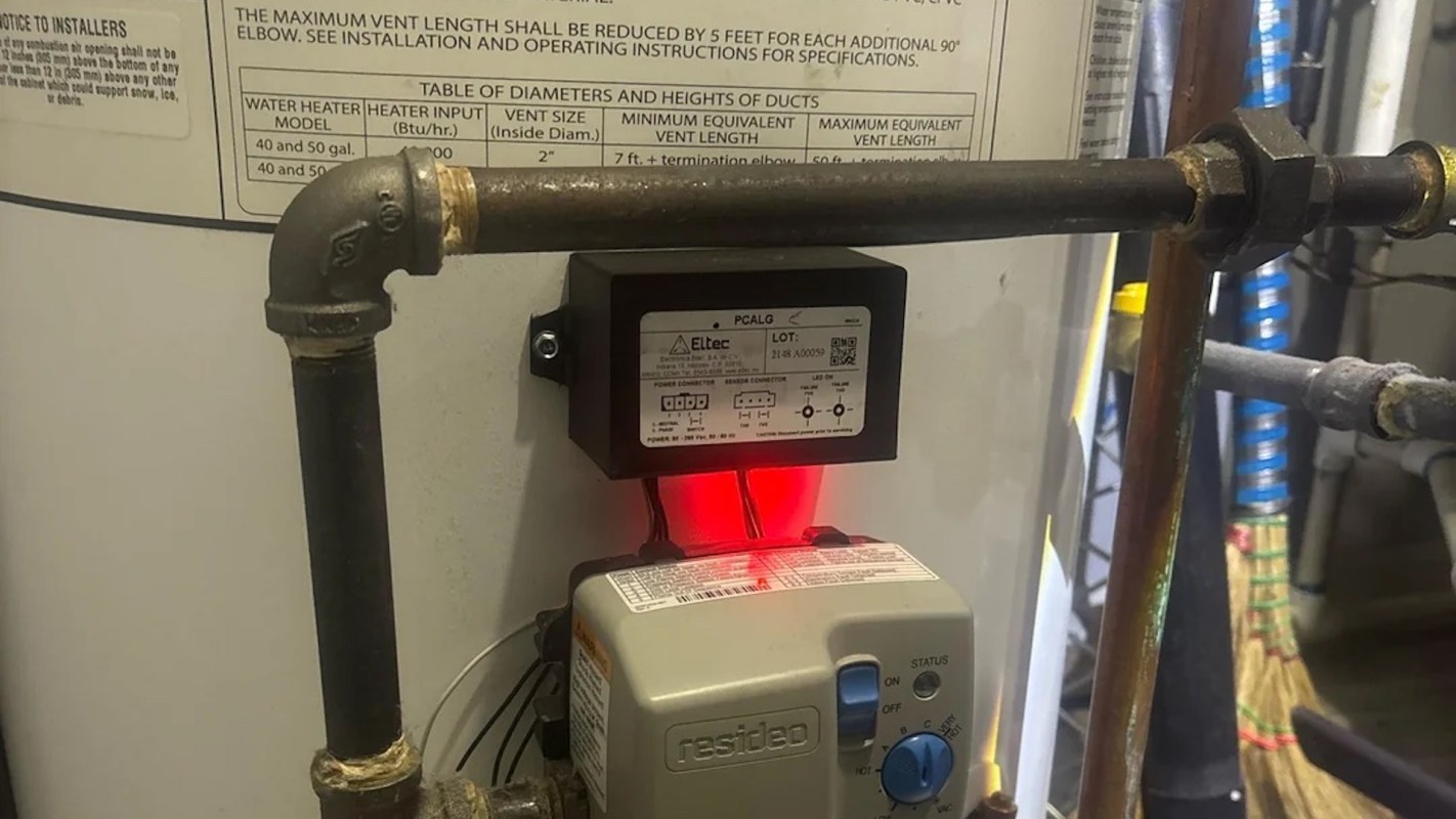 A water heater with connected piping and a control box displaying a red indicator light.
