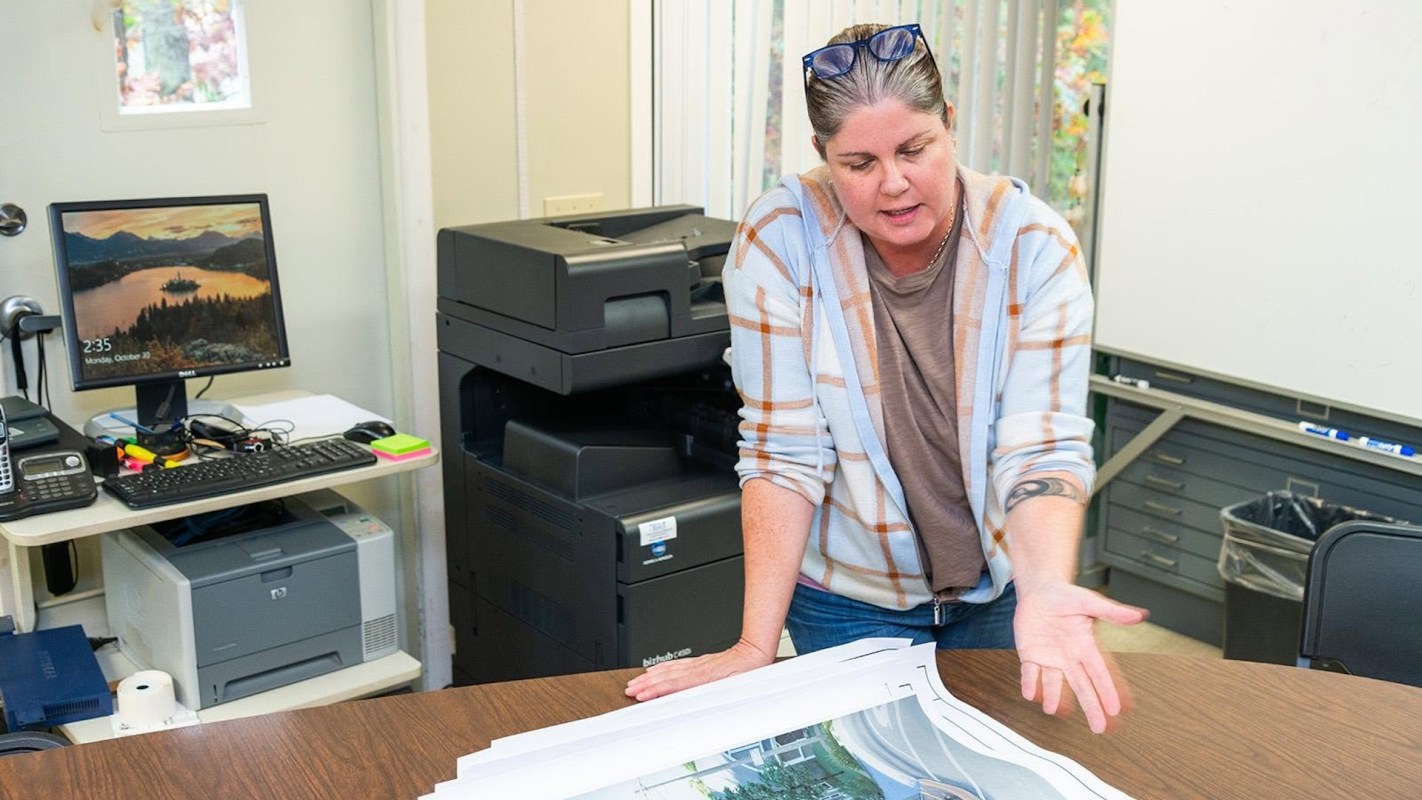 A woman discusses printed plans on a table in an office setting with a computer and a printer in the background.
