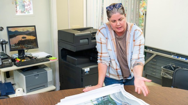 A woman discusses printed plans on a table in an office setting with a computer and a printer in the background.