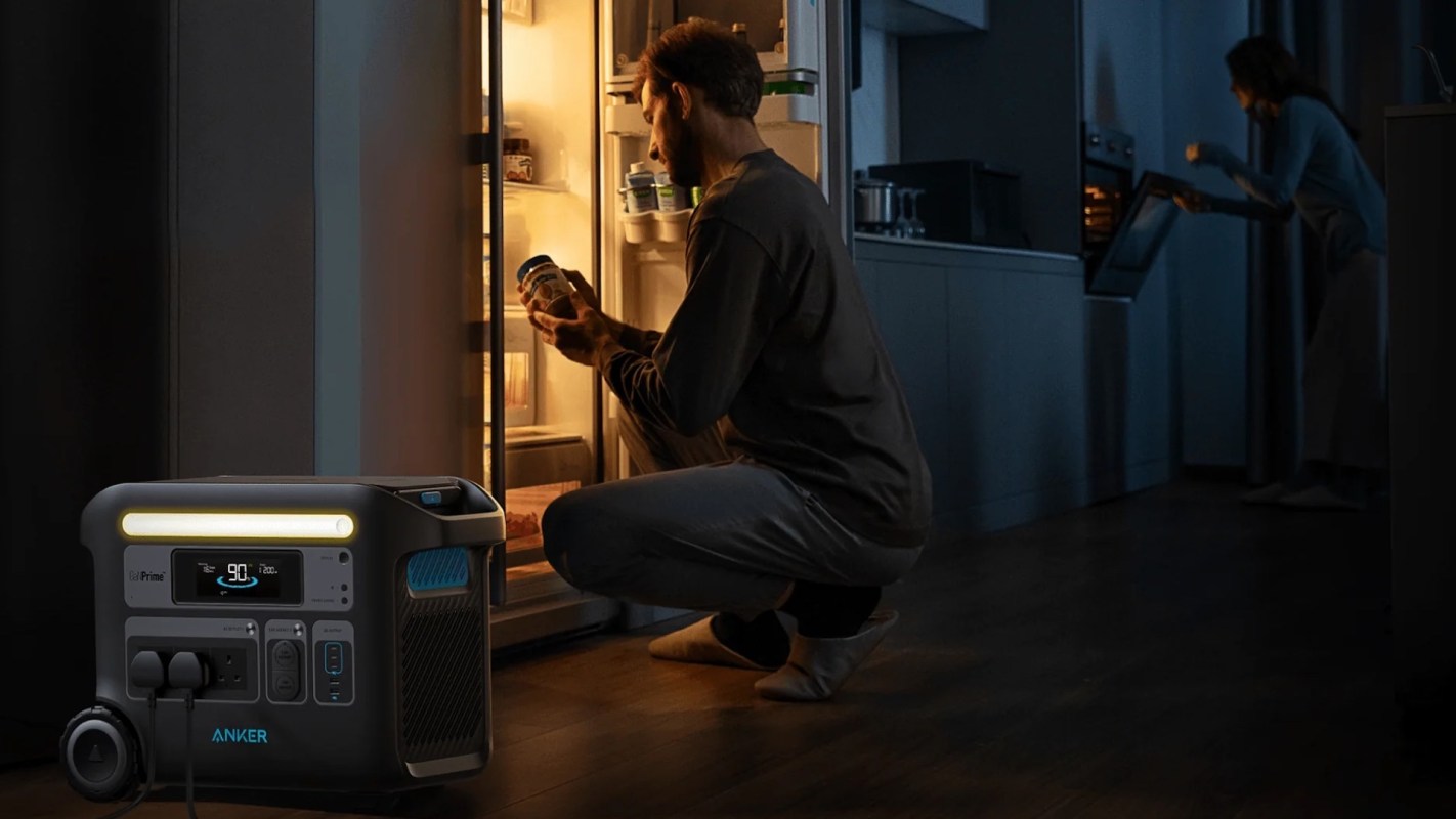 A person kneels in front of an open refrigerator, holding a jar, while a second person looks into an oven.