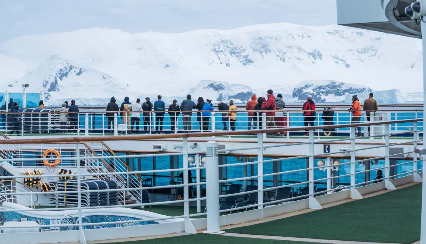 People on a cruise ship deck observe icy Antarctic mountains in the distance.