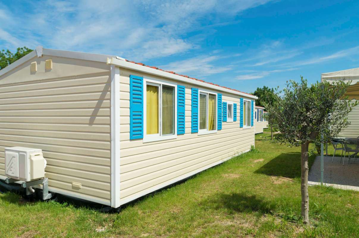 A mobile home with blue shutters and a small tree in a grassy area under a clear blue sky.