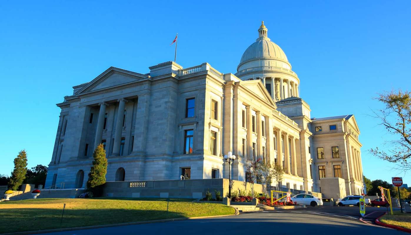 A stone government building with a dome and flag, surrounded by greenery and walkways.