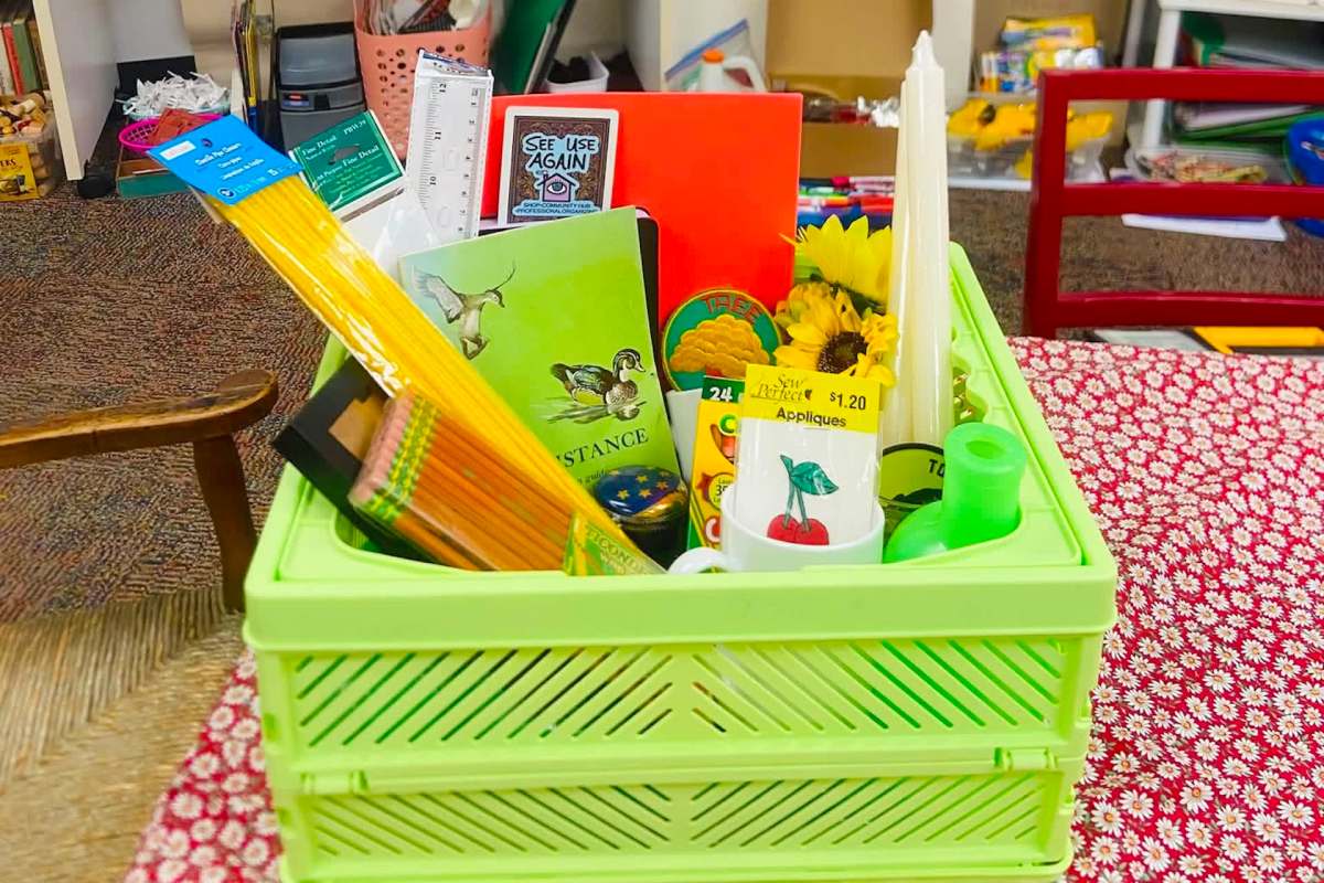 A green basket filled with various school art supplies, including pencils, stickers, flowers, and notebooks.