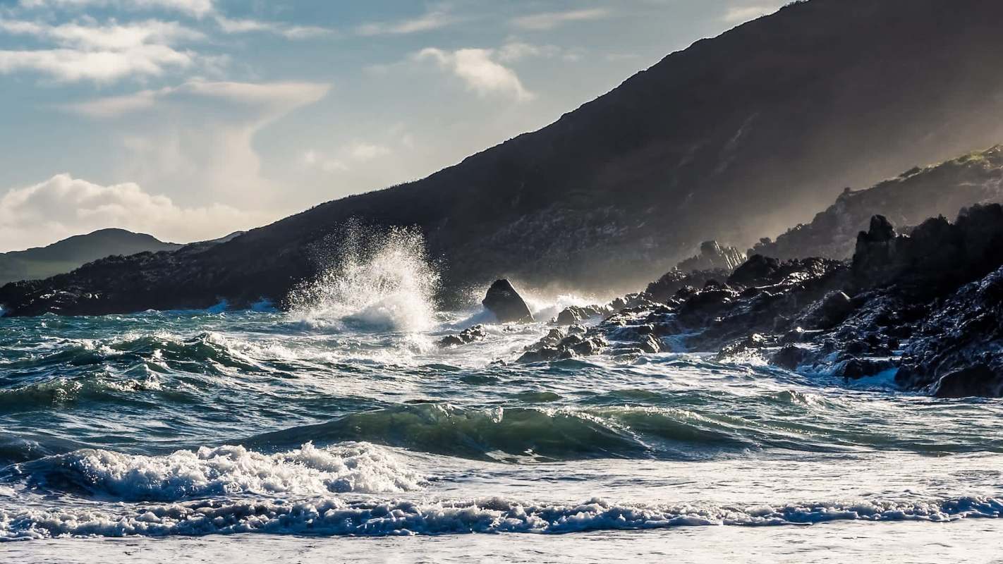 Waves crash against rocky shoreline with mountains in the background under a partly cloudy sky.